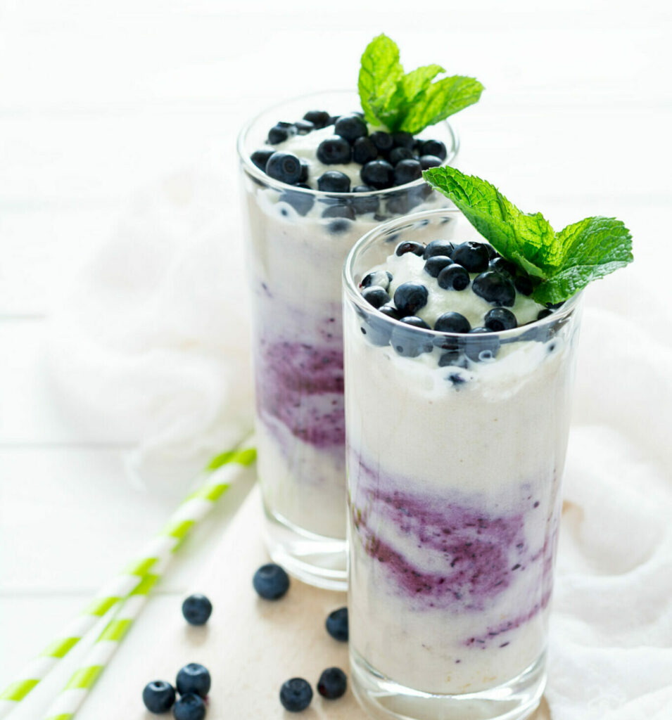 Two Glasses With Fresh Blueberry Smoothie Decorated Berries And Mint Leaves On White Wooden Background. Selective Focus