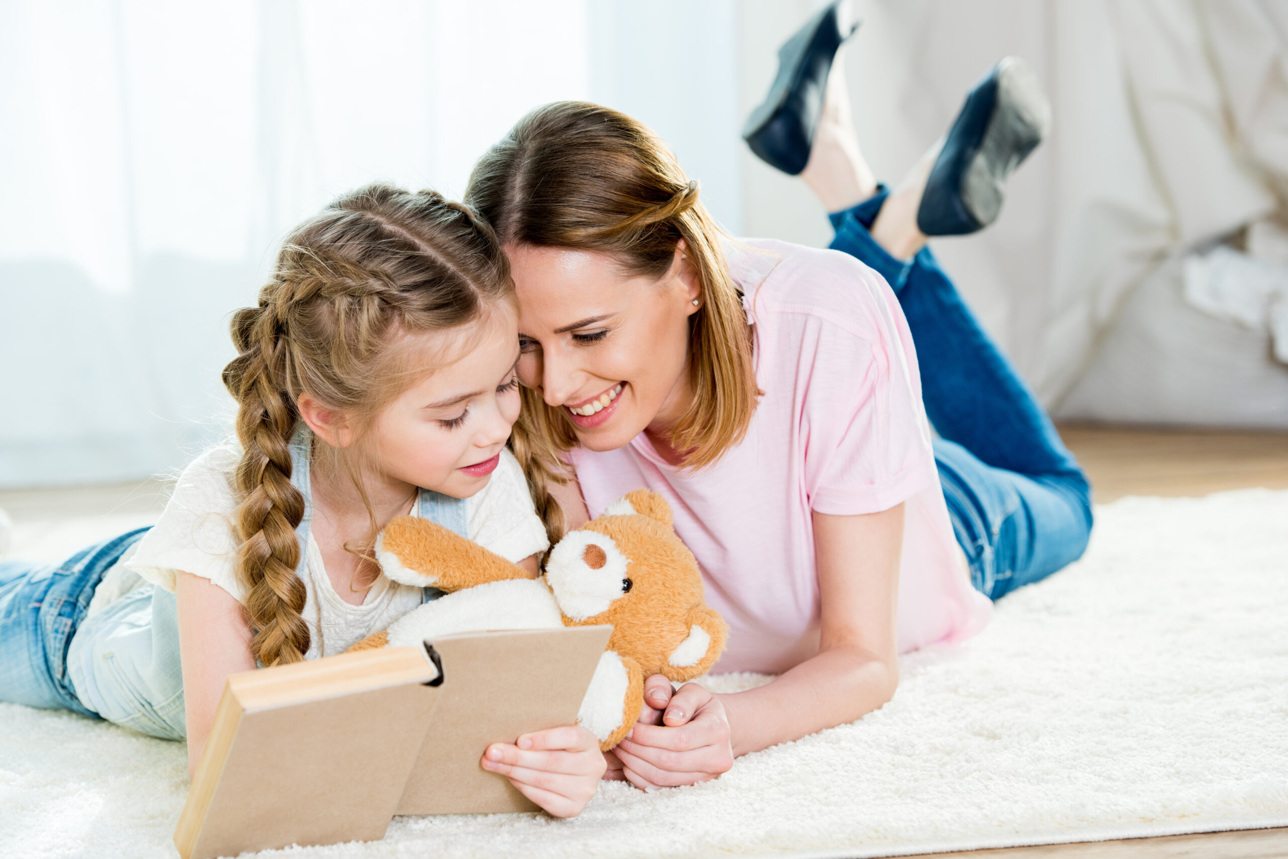 Happy mother and daughter with teddy bear reading book on carpet